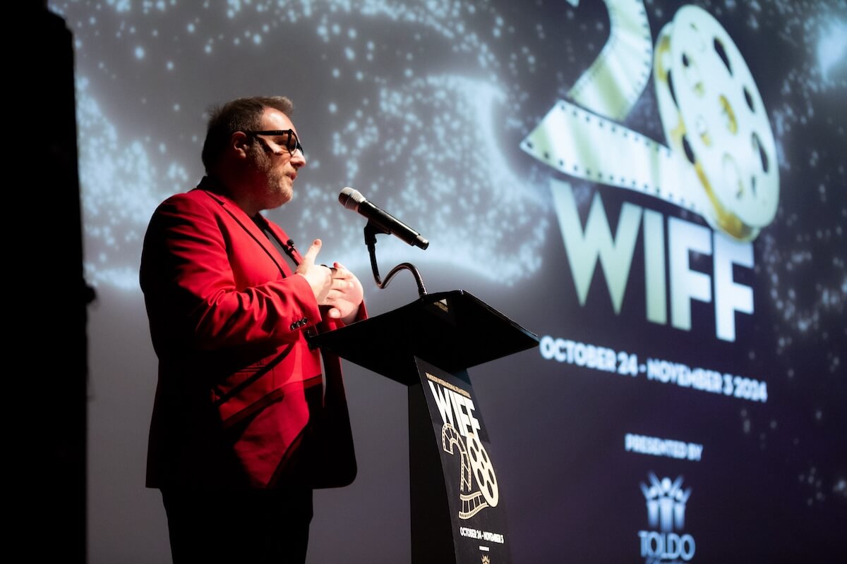 Man speaking at Windsor International Film Festival (WIFF) event. Red suit, podium, film reel logo.